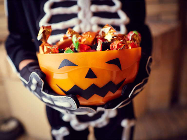 A person dressed in a skeleton costume holds an orange pumpkin-shaped bowl filled with various wrapped candies. The bowl has a classic jack-o'-lantern face with a wide, jagged-toothed smile and triangular eyes and nose. The background is slightly blurred, highlighting the focus on the bowl and its contents, evoking a festive Halloween atmosphere.