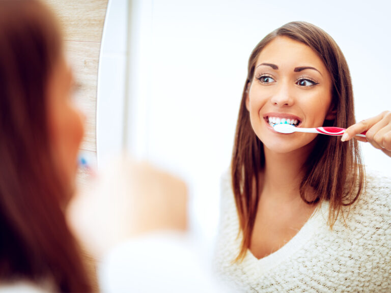 A young woman with long brown hair is brushing her teeth with a red and white toothbrush. She is smiling and looking at herself in a mirror. The background is light and blurry, focusing on her face and the brushing action. She is wearing a white sweater, and her reflection is visible in the mirror on the left side of the image. The scene conveys a sense of routine dental care and hygiene.