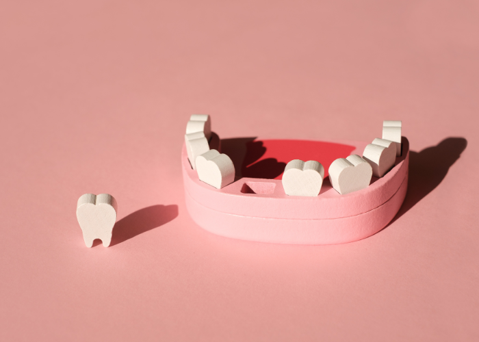 A model of a human lower jaw with artificial teeth placed in a semi-circular pink base. One tooth is positioned outside the model on the left side. The background is a solid light pink, providing a soft and clean look. The setup is well-lit, casting soft shadows and highlighting the details of the artificial teeth and the jaw model.