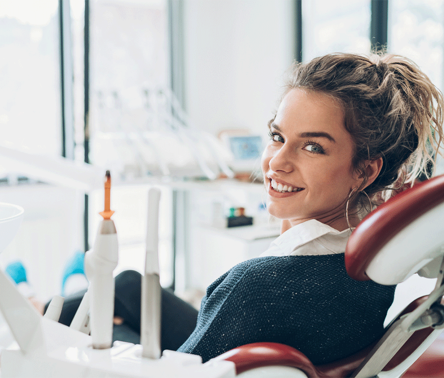 A young woman sitting in a dental chair, smiling at the camera. She has her hair tied back in a ponytail and is wearing a dark sweater over a white shirt. The dental office background shows modern equipment and a bright, clean environment with large windows providing natural light. The woman appears comfortable and happy, indicating a positive dental visit experience.