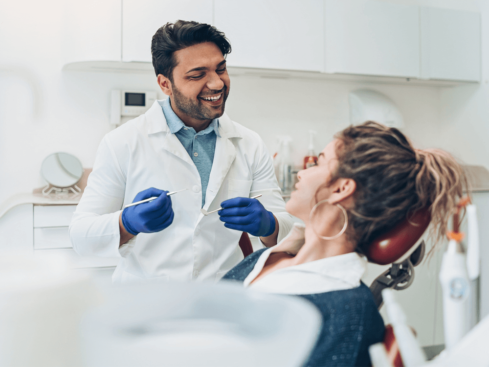 A dentist and a patient in a modern dental office. The dentist, wearing a white coat and blue gloves, is smiling and holding dental instruments while conversing with the patient. The patient, sitting in the dental chair, is also smiling and looking at the dentist. The background shows clean and organized dental equipment and cabinets, creating a professional and welcoming atmosphere.