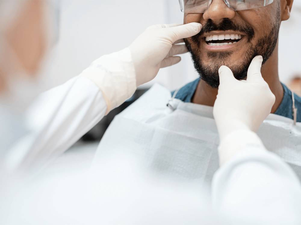 A patient in a dental office receiving a check-up. The patient is smiling and wearing a protective bib, while a dentist, wearing gloves and protective eyewear, gently examines the patient's mouth, focusing on the teeth and gums. The image highlights the patient’s bright smile and the careful attention of the dental professional during the examination.